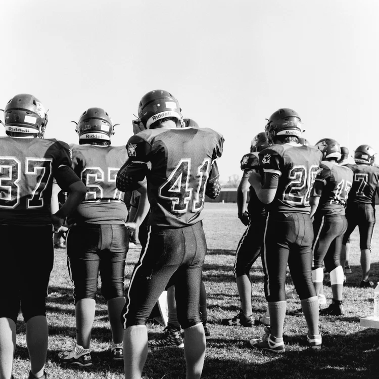 A black-and-white frame of a football team seen from behind on the sideline